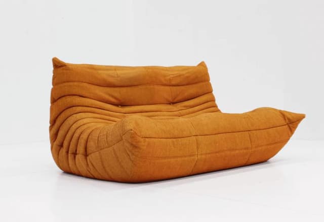 Un canapé moderne, orange et bas, au design doux et ondulé, repose sur un fond blanc. Son allure unique et moelleuse se caractérise par des coutures horizontales incurvées et une atmosphère chaleureuse et accueillante.