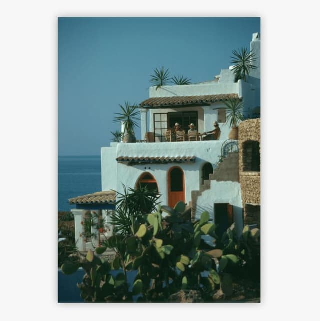 Villa blanche de style méditerranéen surplombant la mer, avec toit en tuiles de terre cuite et fenêtres en arc arrondi. Des palmiers ornent les terrasses. Le ciel bleu éclatant et la mer calme créent une atmosphère sereine.