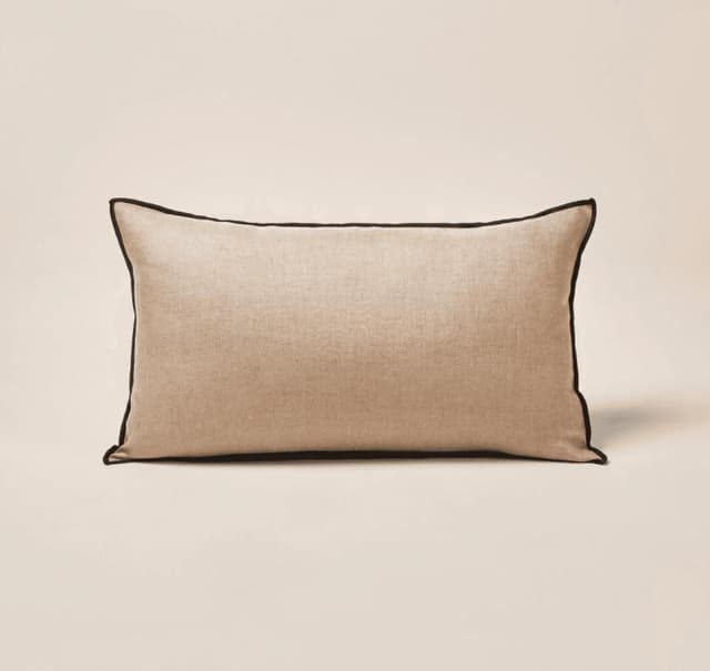 Un coussin rectangulaire beige avec un passepoil noir le long des bords, posé sur un fond neutre.
