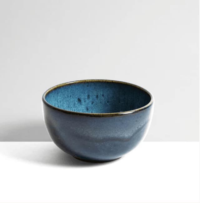 Un bol en céramique à l'extérieur émaillé bleu foncé brillant et à l'intérieur texturé bleu plus clair. Ce bol présente un subtil motif moucheté et repose sur une surface neutre sur un fond gris clair uni.