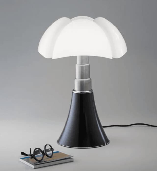 Une lampe de table moderne, dotée d'un abat-jour blanc en forme de pétale et d'une base noire évasée, est posée sur une surface plane, à côté d'une paire de lunettes rondes noires et d'un carnet fermé. Le fond est gris clair uni.