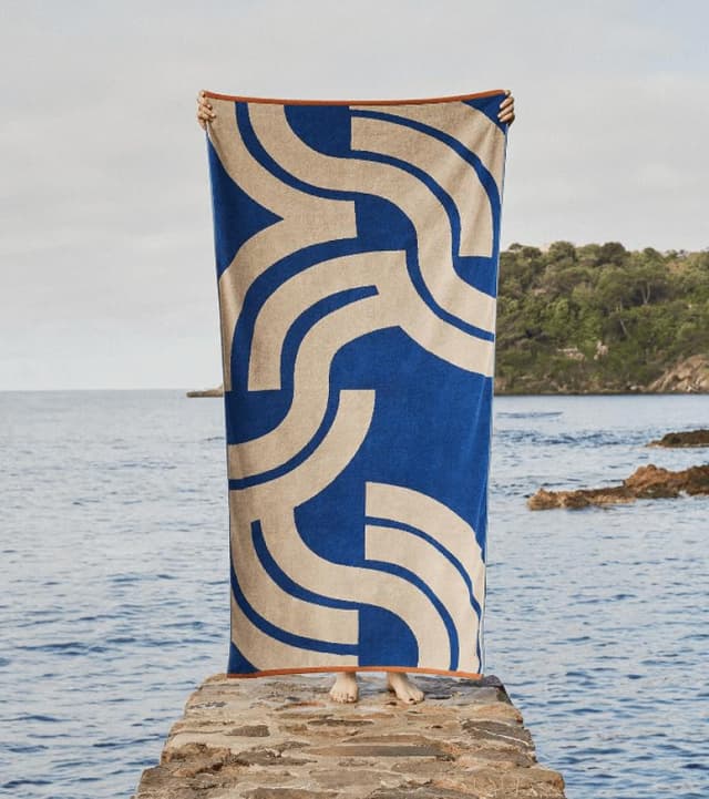 Une personne se tient debout sur une jetée en pierre au bord de la mer, tenant une grande serviette bleue aux motifs géométriques crème audacieux, masquant son corps et son visage. Des arbres et le littoral sont visibles en arrière-plan sous un ciel partiellement nuageux.