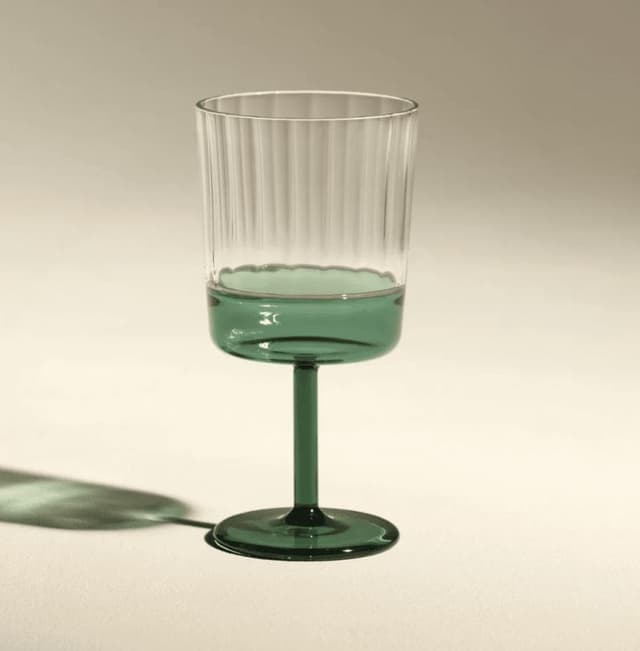 Un gobelet en verre transparent avec des crêtes verticales, comportant une base, une tige et un bol inférieur teintés de vert, posé sur une surface beige clair avec une ombre douce à gauche.