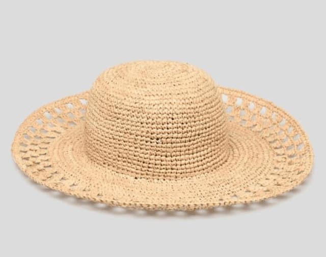 Un chapeau de paille tressé à large bord avec un design ouvert et aéré autour du bord, placé sur un fond gris clair uni.