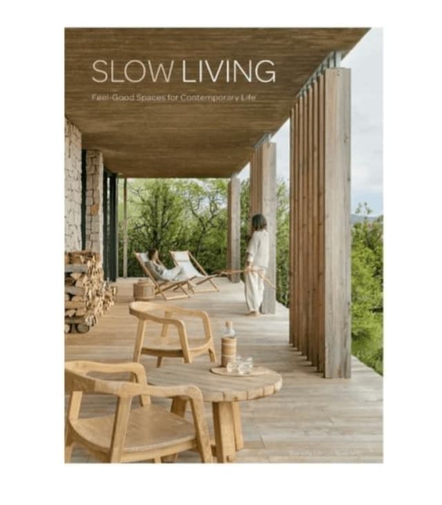 Une terrasse moderne en bois avec des chaises longues et une table surplombe la verdure. Une personne en tenue légère se tient près de la balustrade et regarde dehors. La scène est paisible, avec les mots "Slow Living" écrits en haut.
