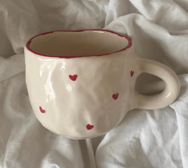 Une tasse en céramique de couleur crème avec un bord rouge et des petits cœurs rouges est posée sur un fond de tissu blanc. Le mug a un aspect légèrement irrégulier, fait à la main.