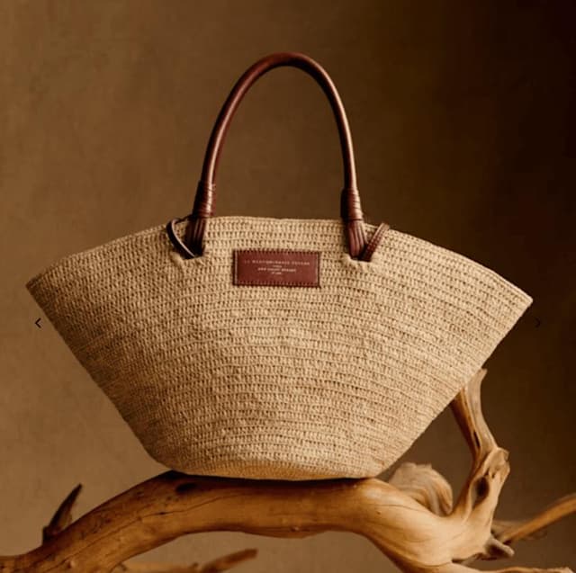 Un sac fourre-tout en paille tressée avec deux poignées incurvées en cuir marron et un écusson rectangulaire en cuir marron sur le devant, présenté sur un morceau de bois flotté sur un fond marron.