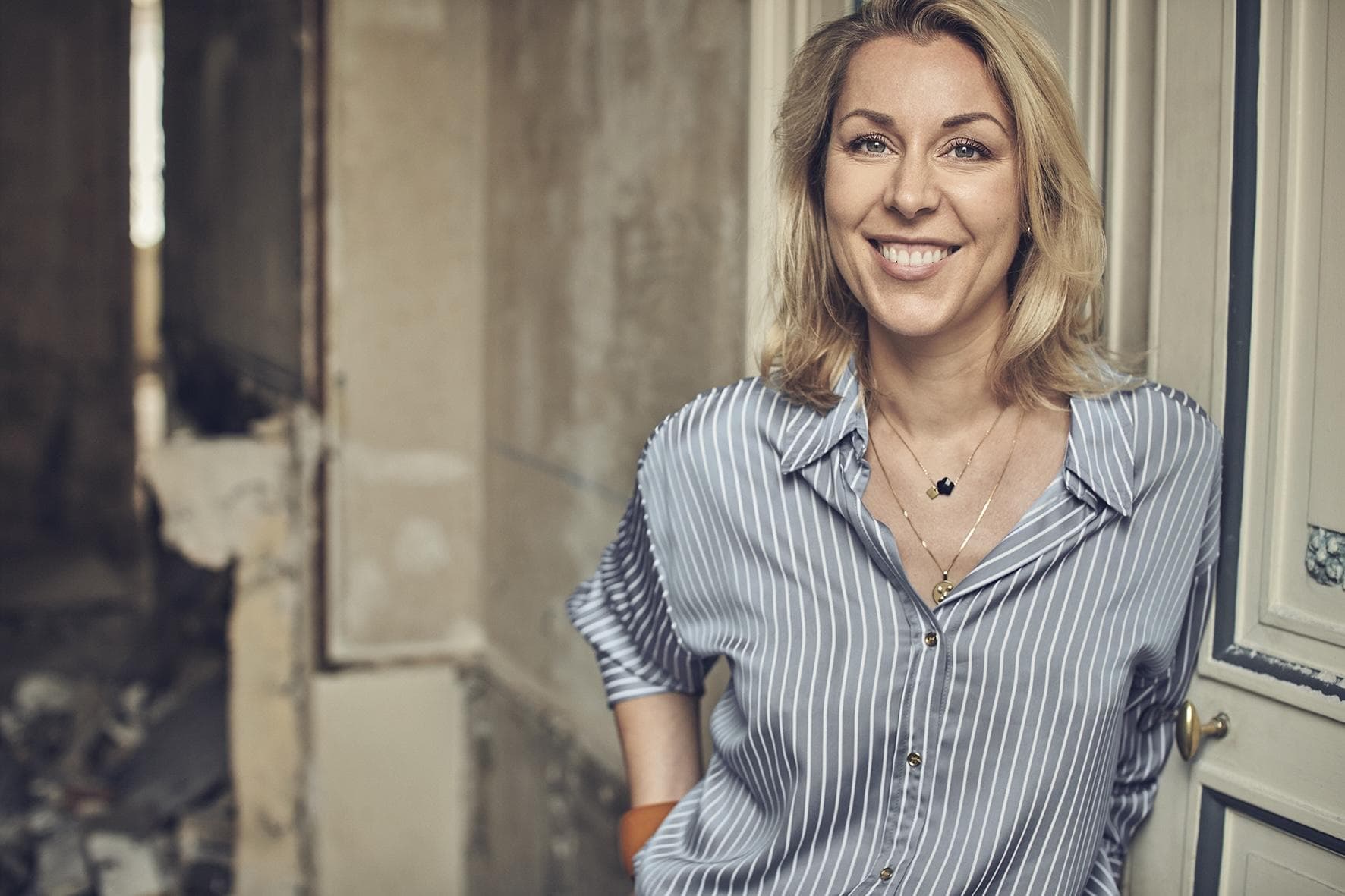 A woman with blonde hair wearing a blue and white striped shirt smiles while standing by a doorway in a partially renovated or unfinished room.