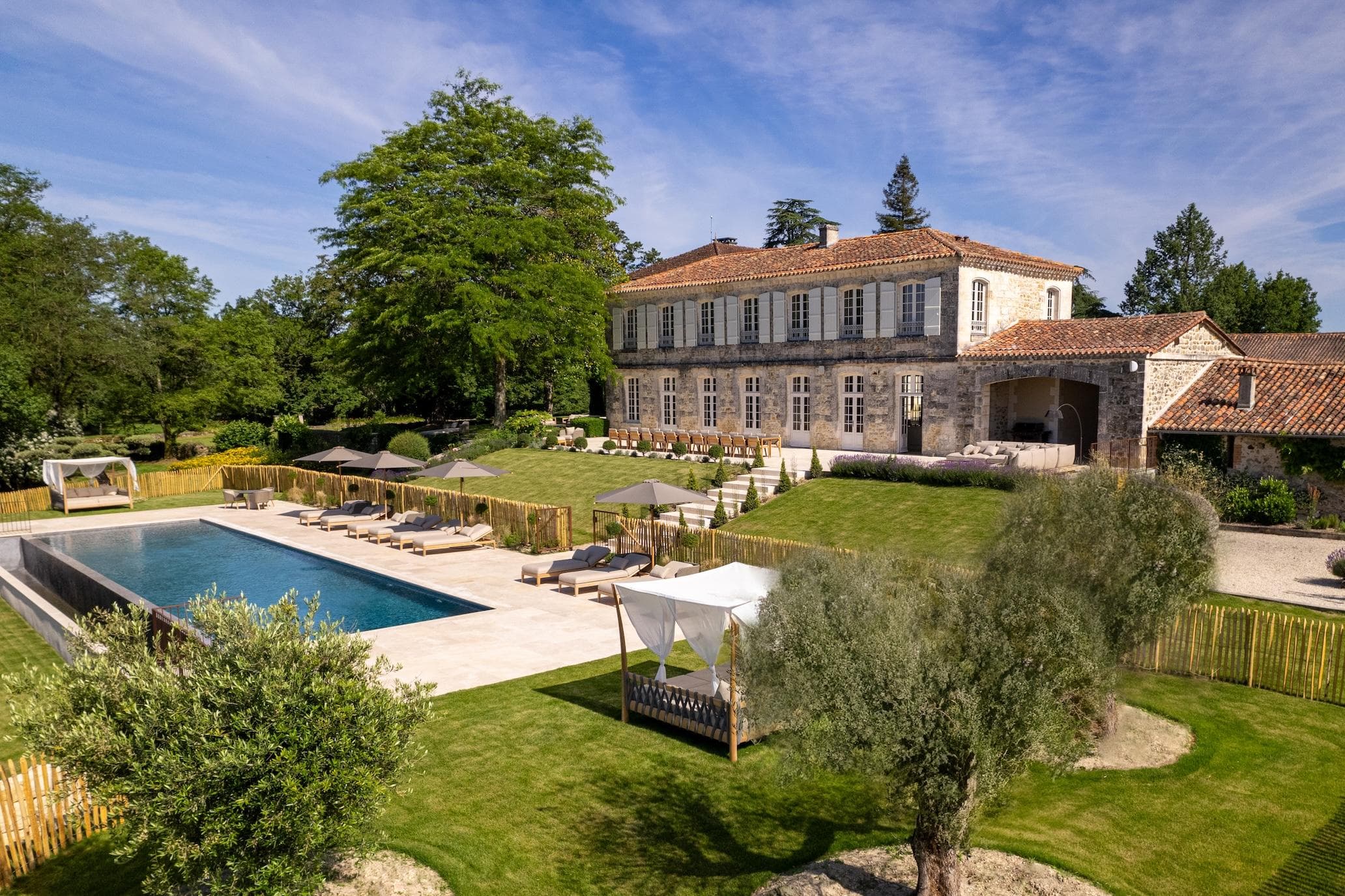 A luxurious stone villa with red-tiled roof sits on a grassy hill, overlooking a large rectangular swimming pool with lounge chairs and umbrellas, surrounded by lush greenery and tall trees under a blue sky.