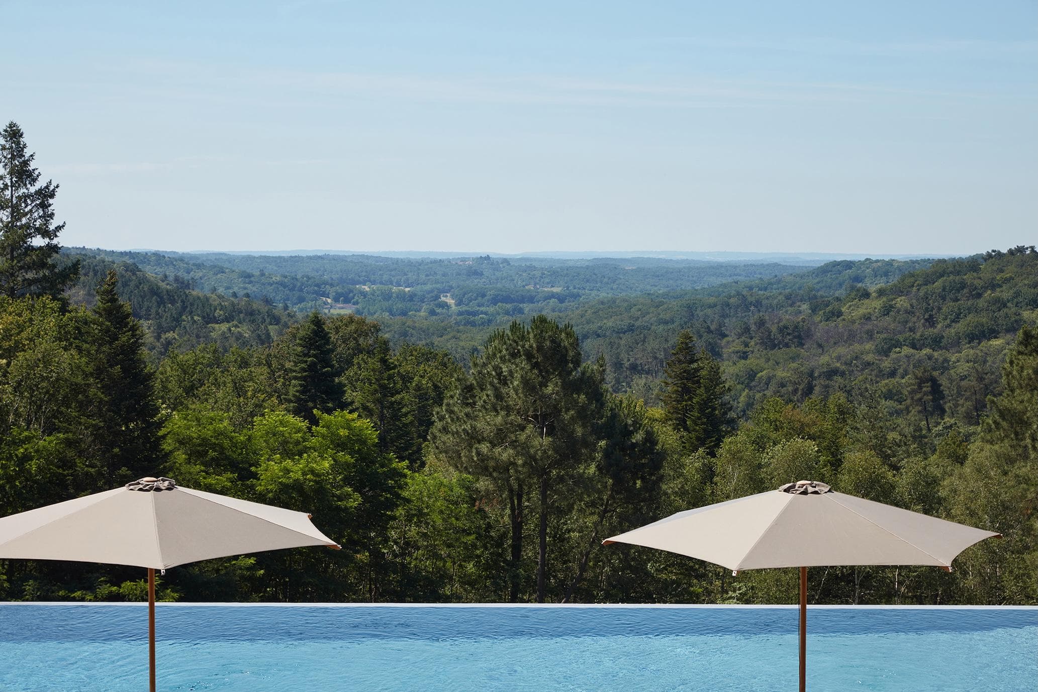 Two white umbrellas stand by the edge of an infinity pool, overlooking a lush green forest with rolling hills and a clear blue sky in the distance.