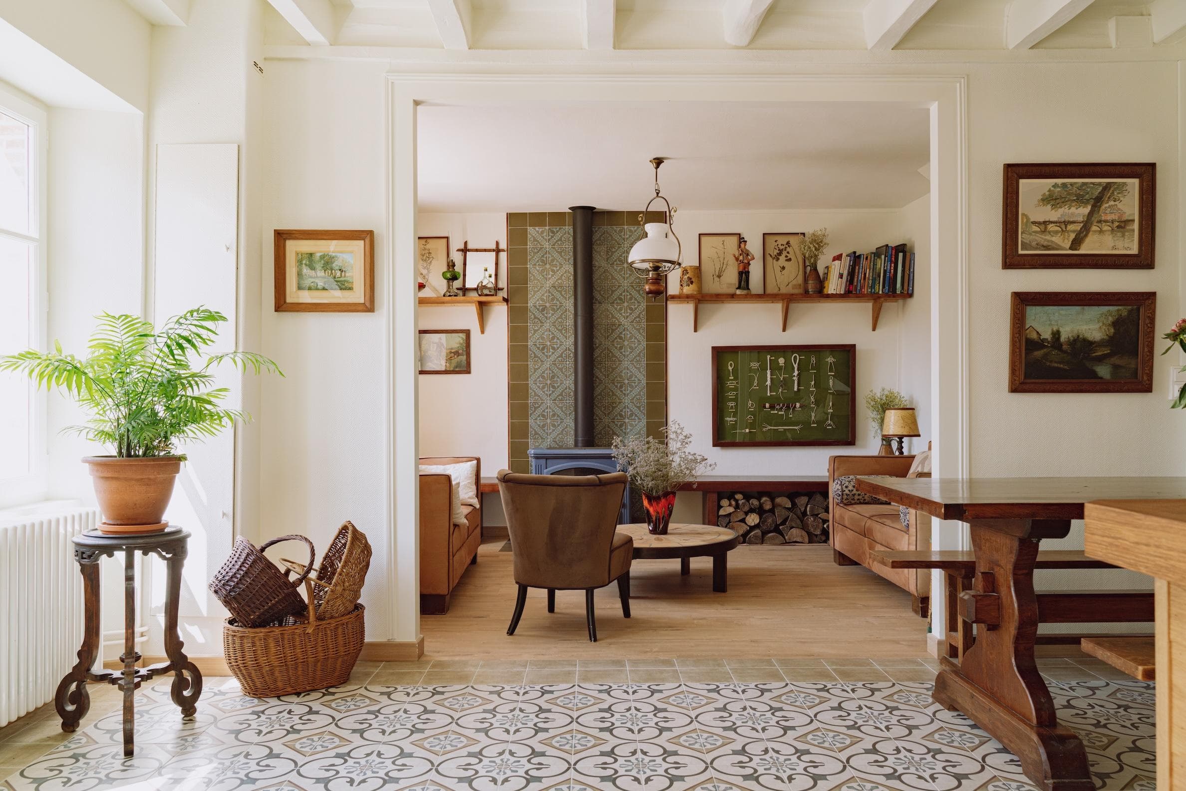 A cozy, sunlit living room with wooden furniture, patterned floor tiles, potted plants, framed art, a wood-burning stove, baskets, and bookshelves. The space feels warm and inviting with natural light streaming in.