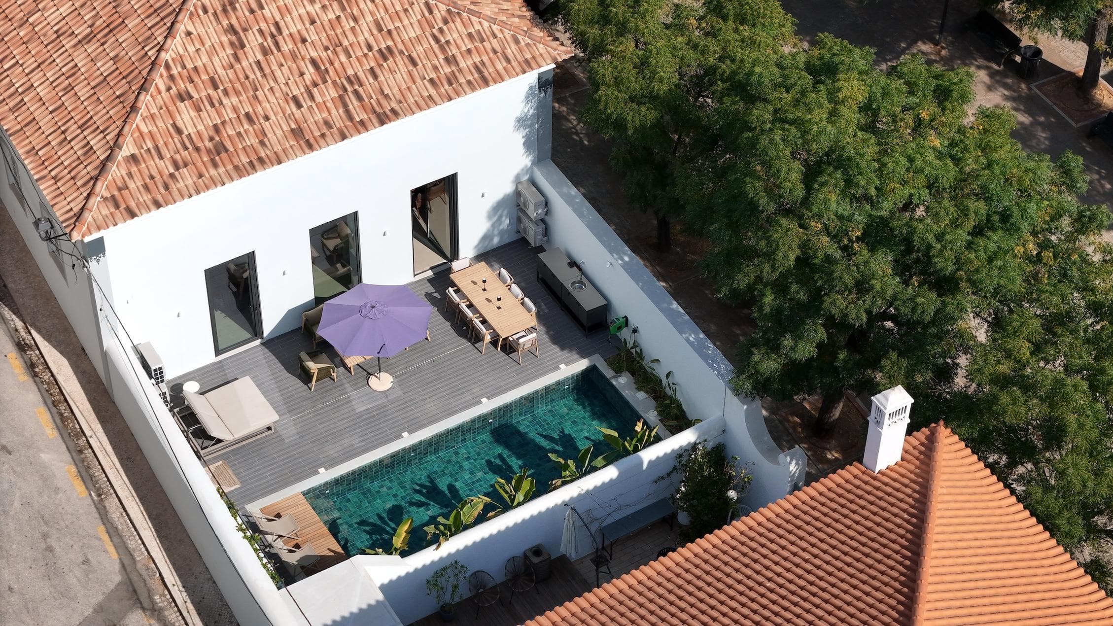 Aerial view of a modern house with a tiled roof, a small rectangular pool, lounge chairs, a dining table, and a purple umbrella on a wooden deck surrounded by white walls and trees.