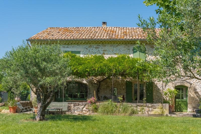 Une maison rustique en pierre avec un toit en tuiles et des volets verts, entourée d'arbres luxuriants, de vignes et d'une cour herbeuse sous un ciel bleu clair. Un patio ombragé avec des plantes et des meubles d'extérieur est visible à l'avant.