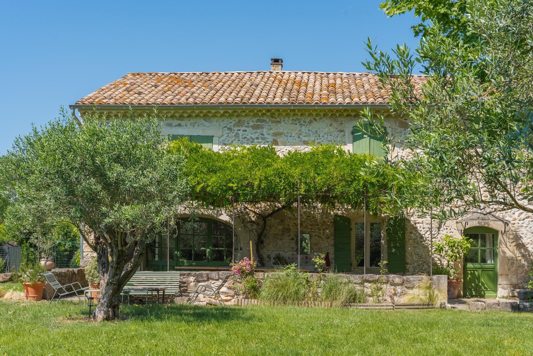 Une maison rustique en pierre avec un toit en tuiles et des volets verts, entourée d'arbres luxuriants, de vignes et d'une cour herbeuse sous un ciel bleu clair. Un patio ombragé avec des plantes et des meubles d'extérieur est visible à l'avant.