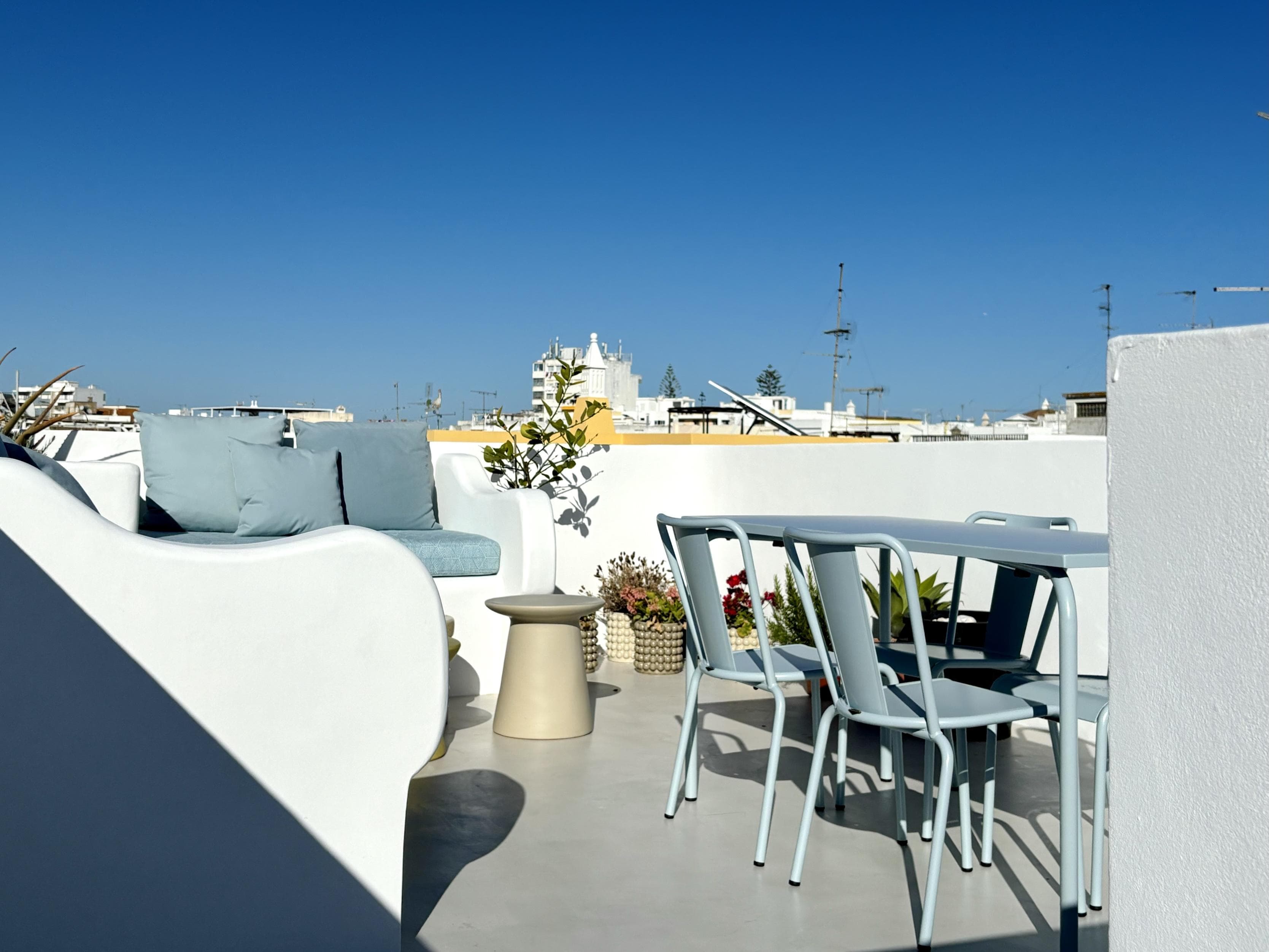 A rooftop terrace with a white sofa, blue cushions, a small beige side table, a blue dining table, matching chairs, and potted plants, all under a clear blue sky with city buildings in the background.