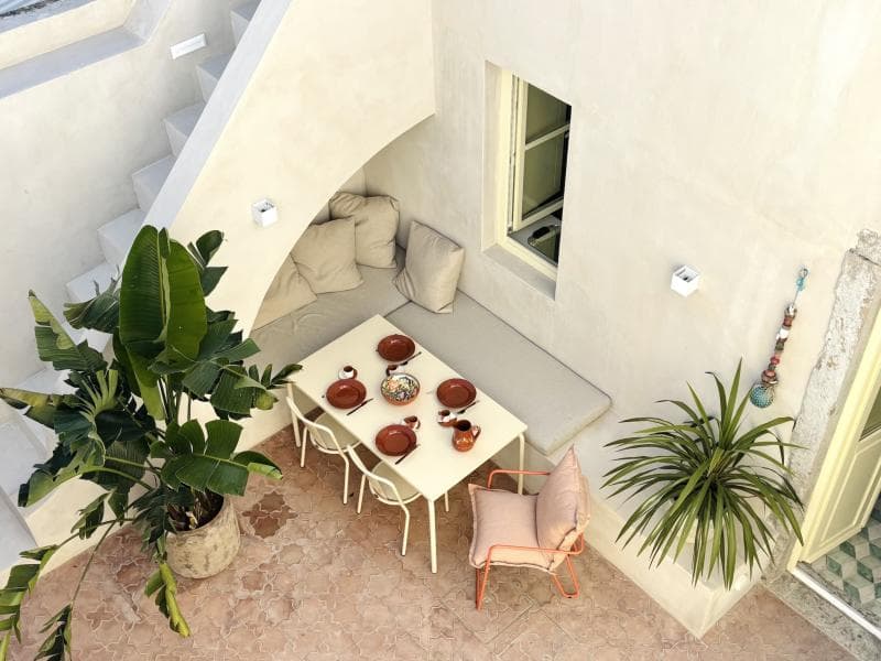 Un patio extérieur confortable avec une petite table à manger pour cinq personnes, entourée de bancs rembourrés, d'une chaise rose, de grandes plantes en pot et de murs blancs avec des escaliers. La lumière du soleil éclaire le sol carrelé et l'espace accueillant.