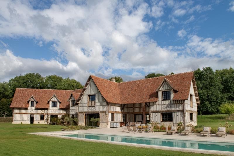A large, traditional half-timbered house with a red-tiled roof sits next to a rectangular swimming pool, surrounded by green lawn and trees under a partly cloudy sky.