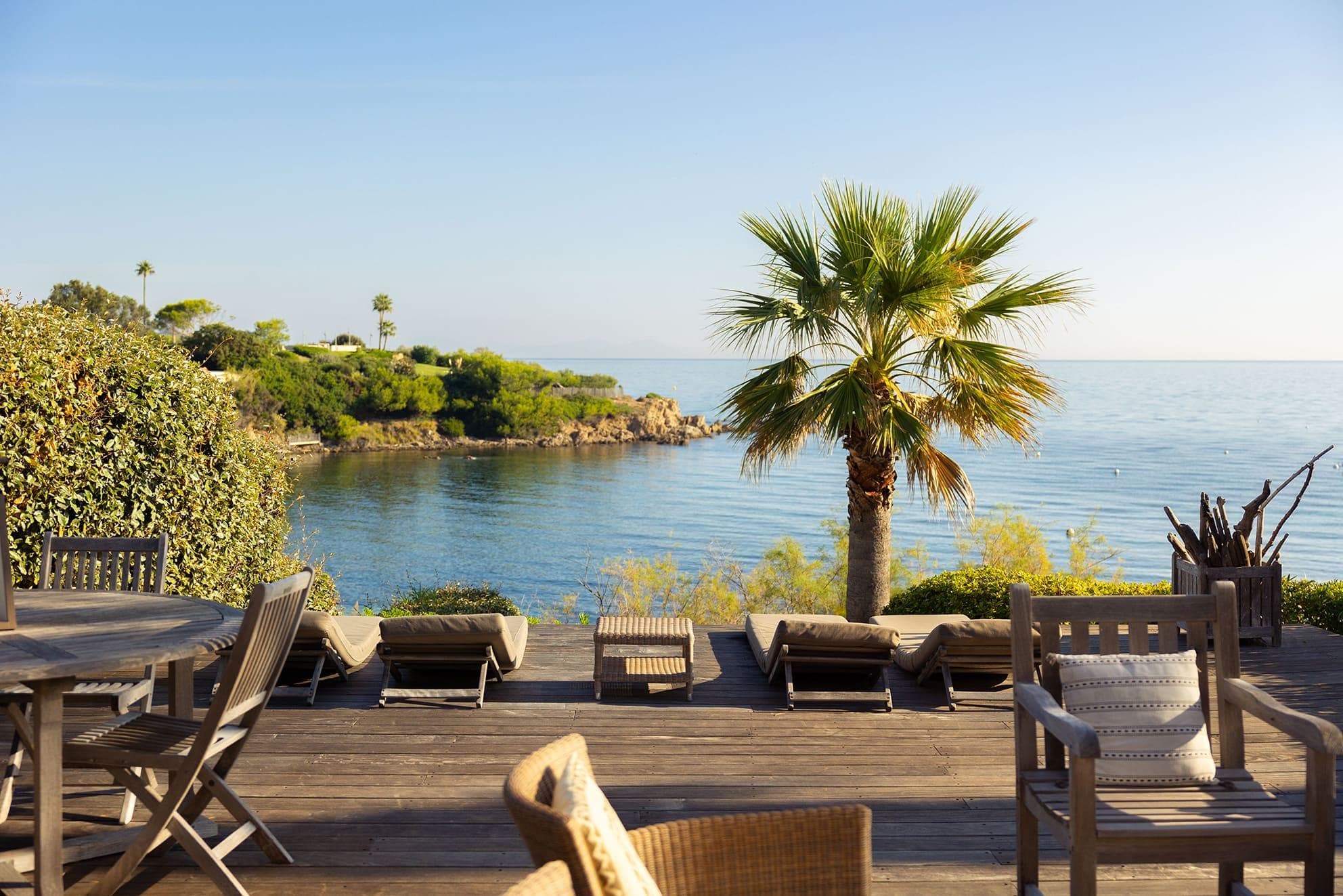 A sunny deck with wooden chairs and tables overlooks the calm sea, with lounge chairs facing the water, a palm tree, and green coastal foliage in the background.