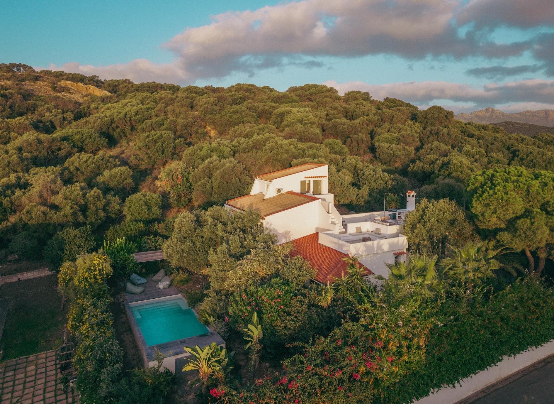 A modern white house with a pool is surrounded by lush green trees and hills under a blue sky with scattered clouds. The scene is tranquil and sunlit.