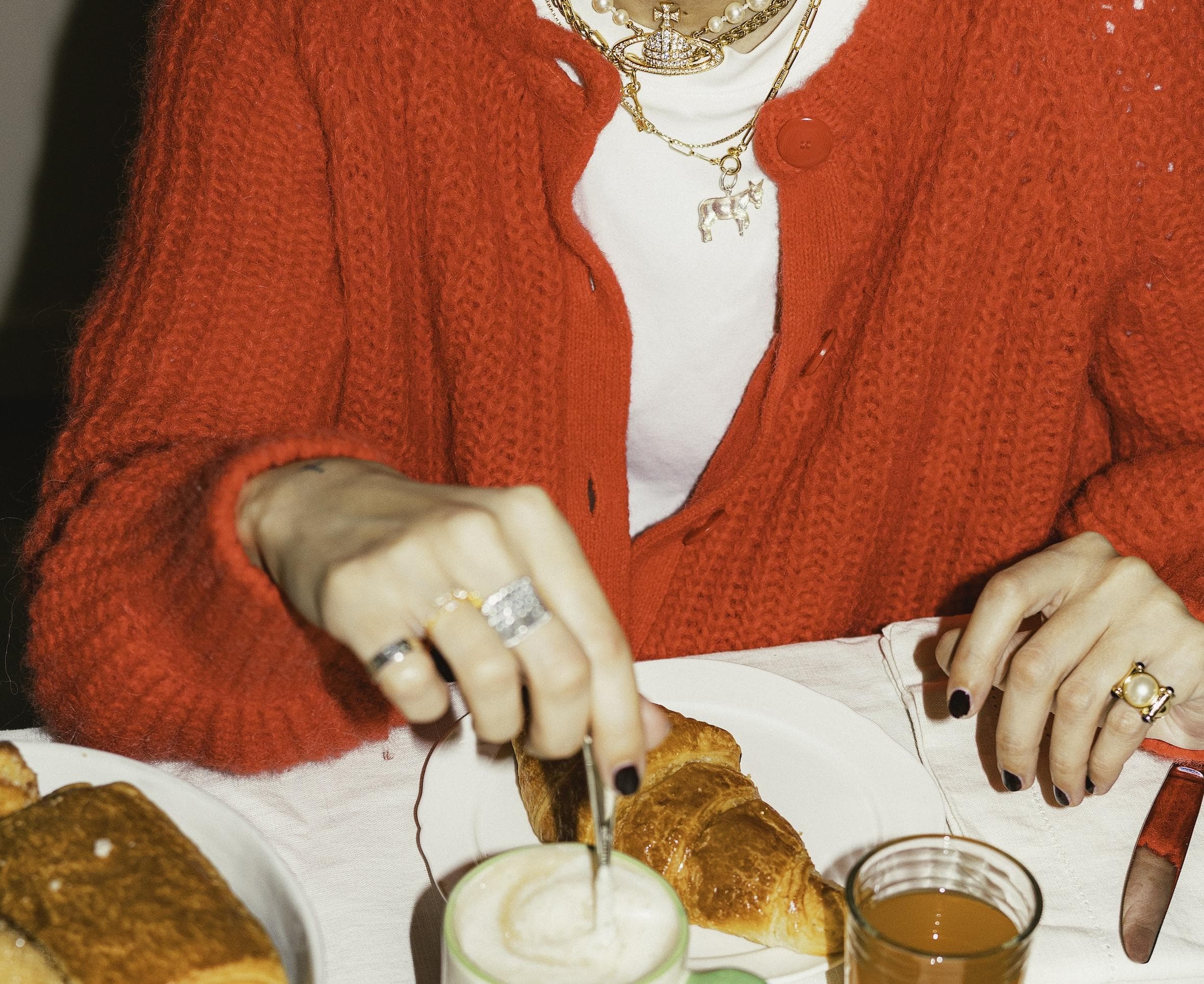 A person wearing a bright red sweater and gold jewelry stirs a foamy drink at a breakfast table with croissants, juice, and a jar of jam. Only their hands and torso are visible.