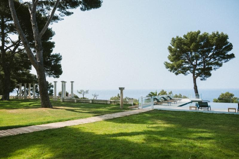 A grassy lawn with a stone path leads to a pool area with lounge chairs, surrounded by trees and classical columns, overlooking the sea under a clear blue sky.