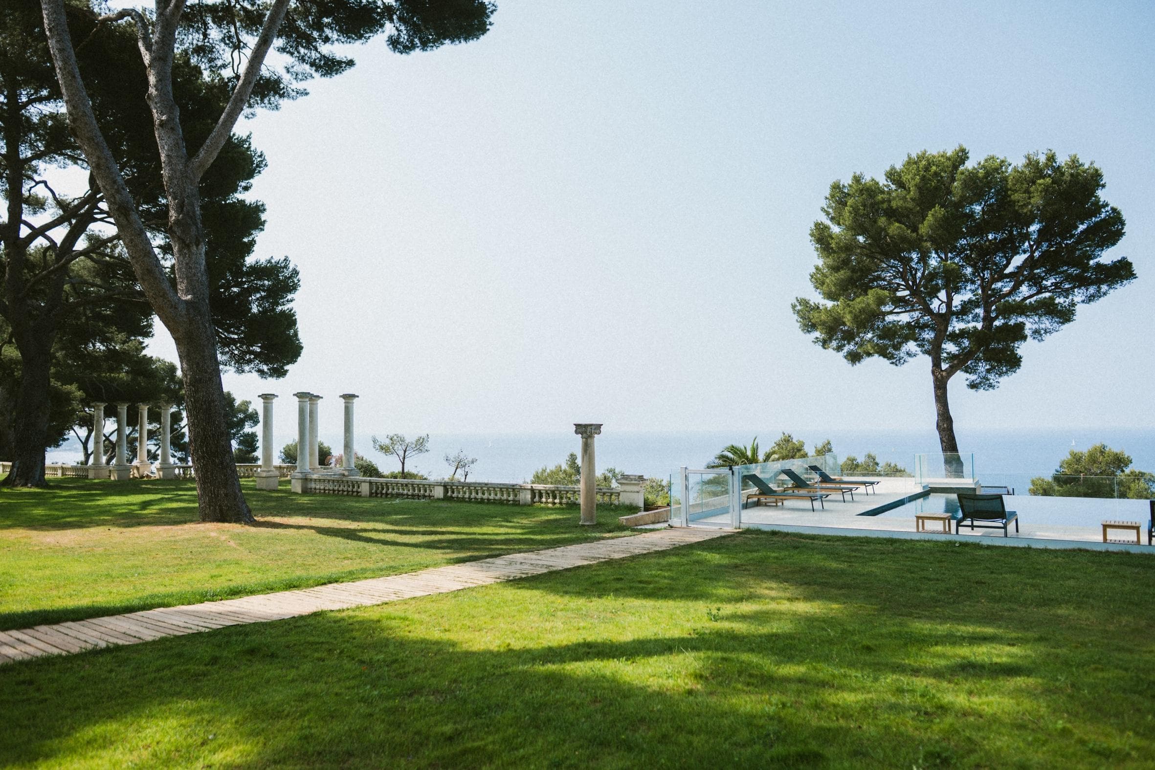 A grassy lawn with a stone path leads to a pool area with lounge chairs, surrounded by trees and classical columns, overlooking the sea under a clear blue sky.