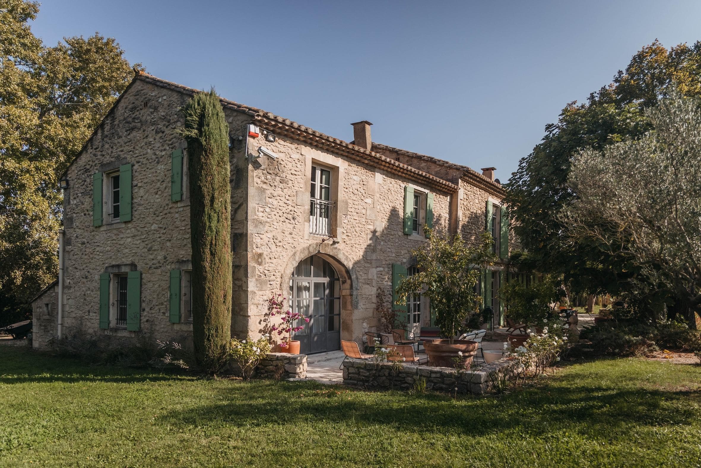 A rustic stone house with green shutters and a tiled roof, surrounded by trees and a green lawn, basking in warm sunlight. A small balcony and potted plants decorate the front entrance.