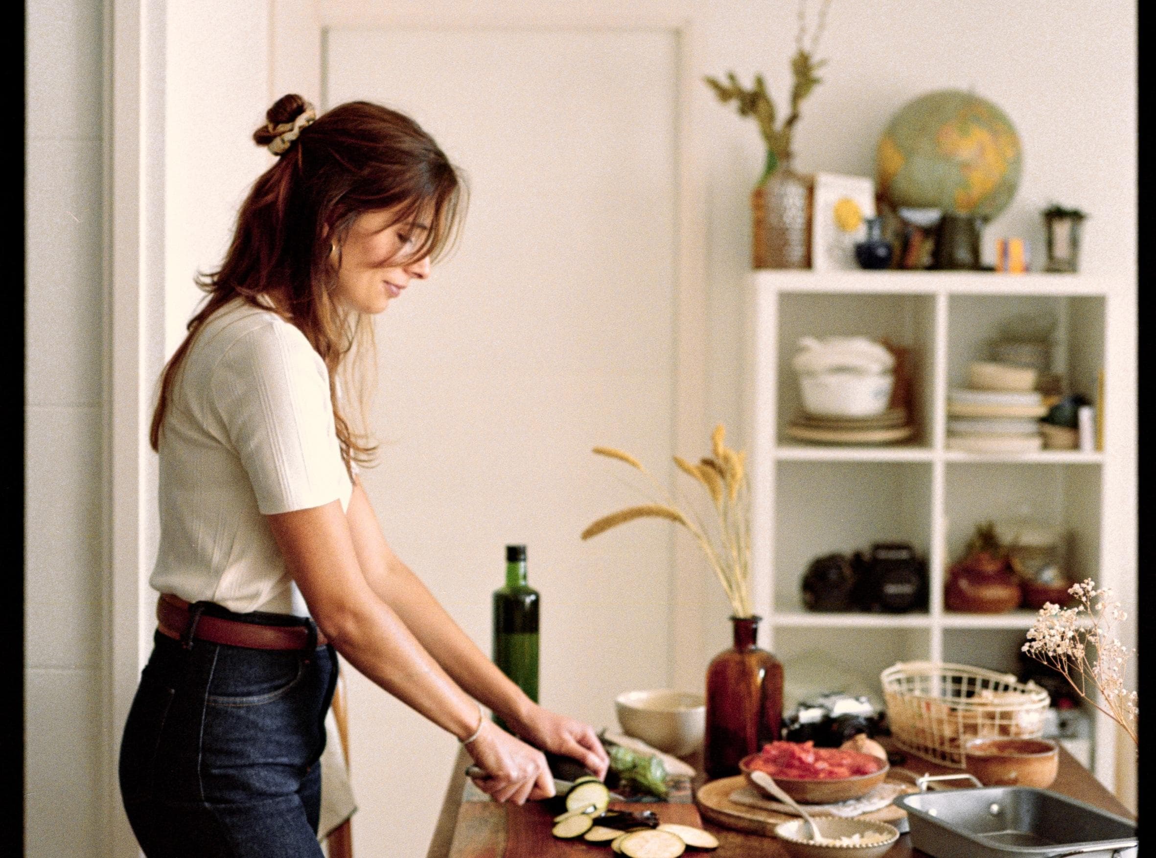 A woman with long hair tied back is chopping vegetables in a cozy kitchen. Shelves with dishes, plants, and a globe are visible in the background. A bottle of olive oil and fresh ingredients are on the counter.