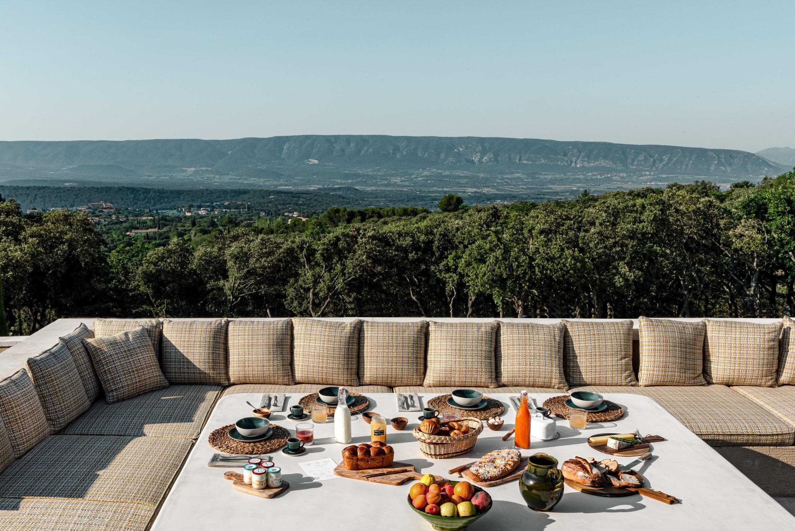 Une configuration de salle à manger en plein air comprenant une grande table carrée avec des sièges rembourrés beiges. La table est ornée d’assiettes, de boissons et de plats variés, sur fond de verdure luxuriante et de montagnes lointaines sous un ciel dégagé.