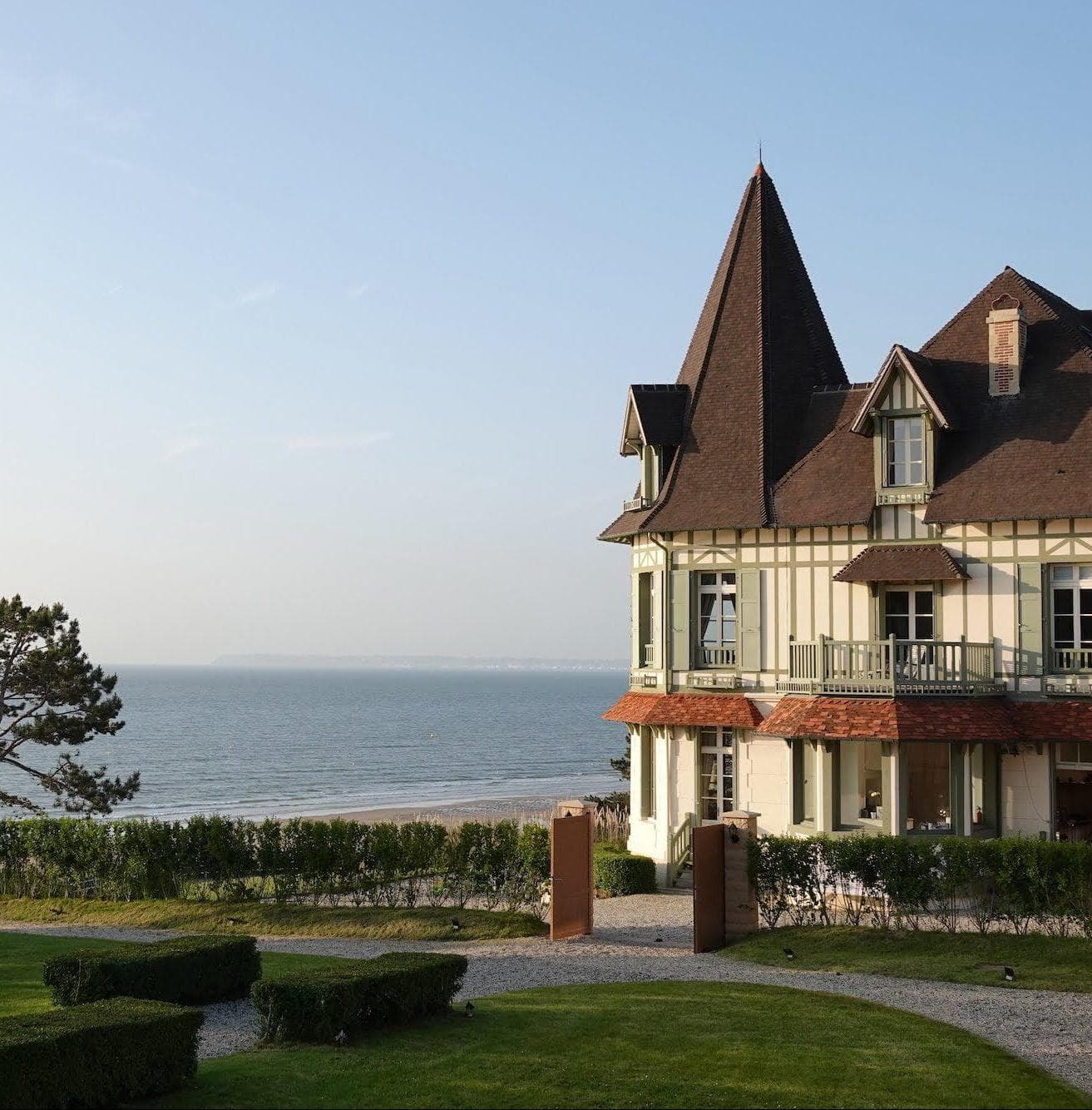 Une belle maison de bord de mer avec une tour pointue et un design à colombages se dresse sur une pelouse verte, surplombant un océan calme. Le ciel bleu clair met en valeur le paysage côtier tranquille, ce qui en fait un lieu de retraite idéal pour les amateurs de yoga en quête de paix et de sérénité.