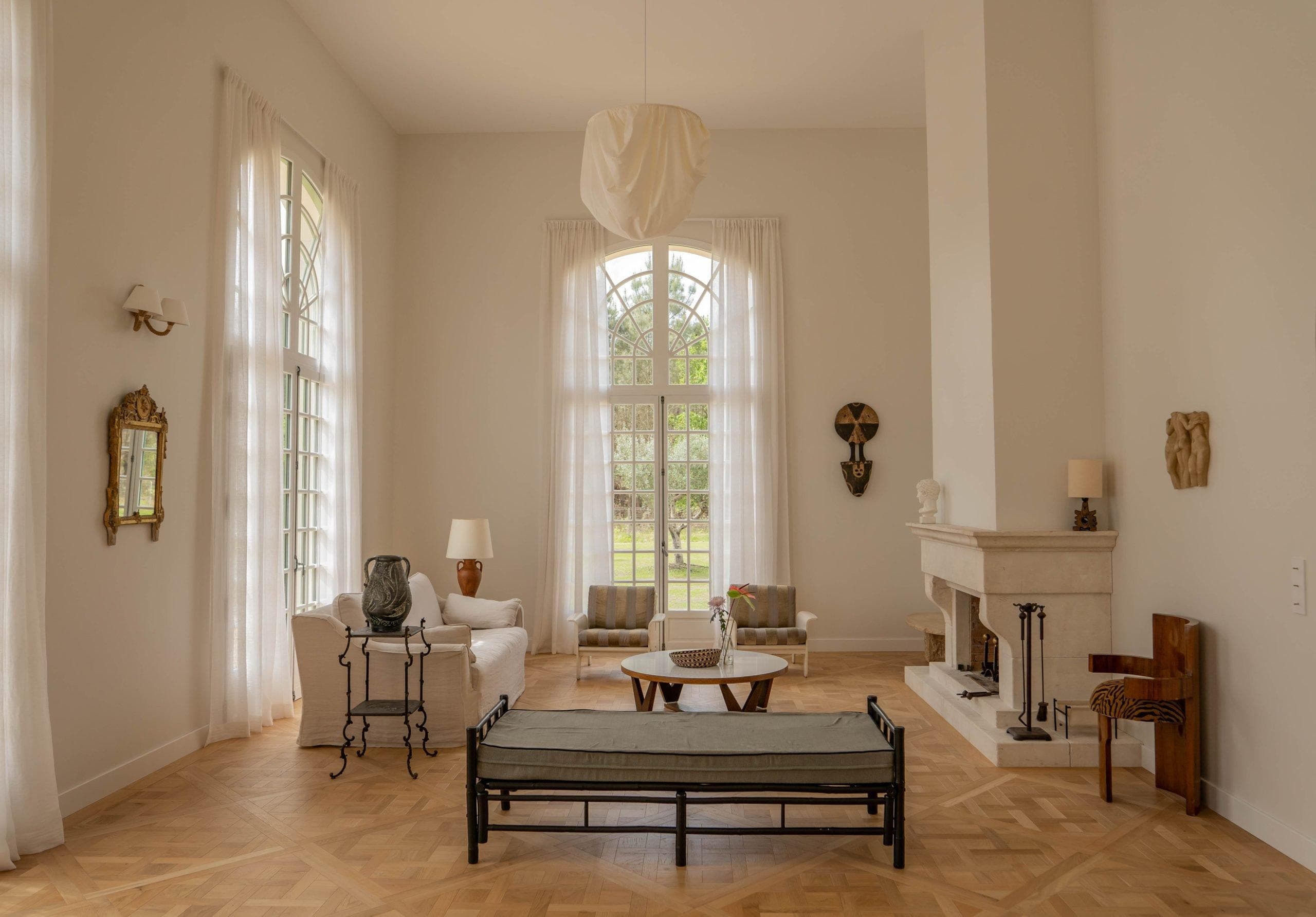 A spacious, elegant living room with tall arched windows, sheer white curtains, neutral-toned furniture, a fireplace, and wooden parquet flooring. Art pieces and decor items are arranged throughout the room.