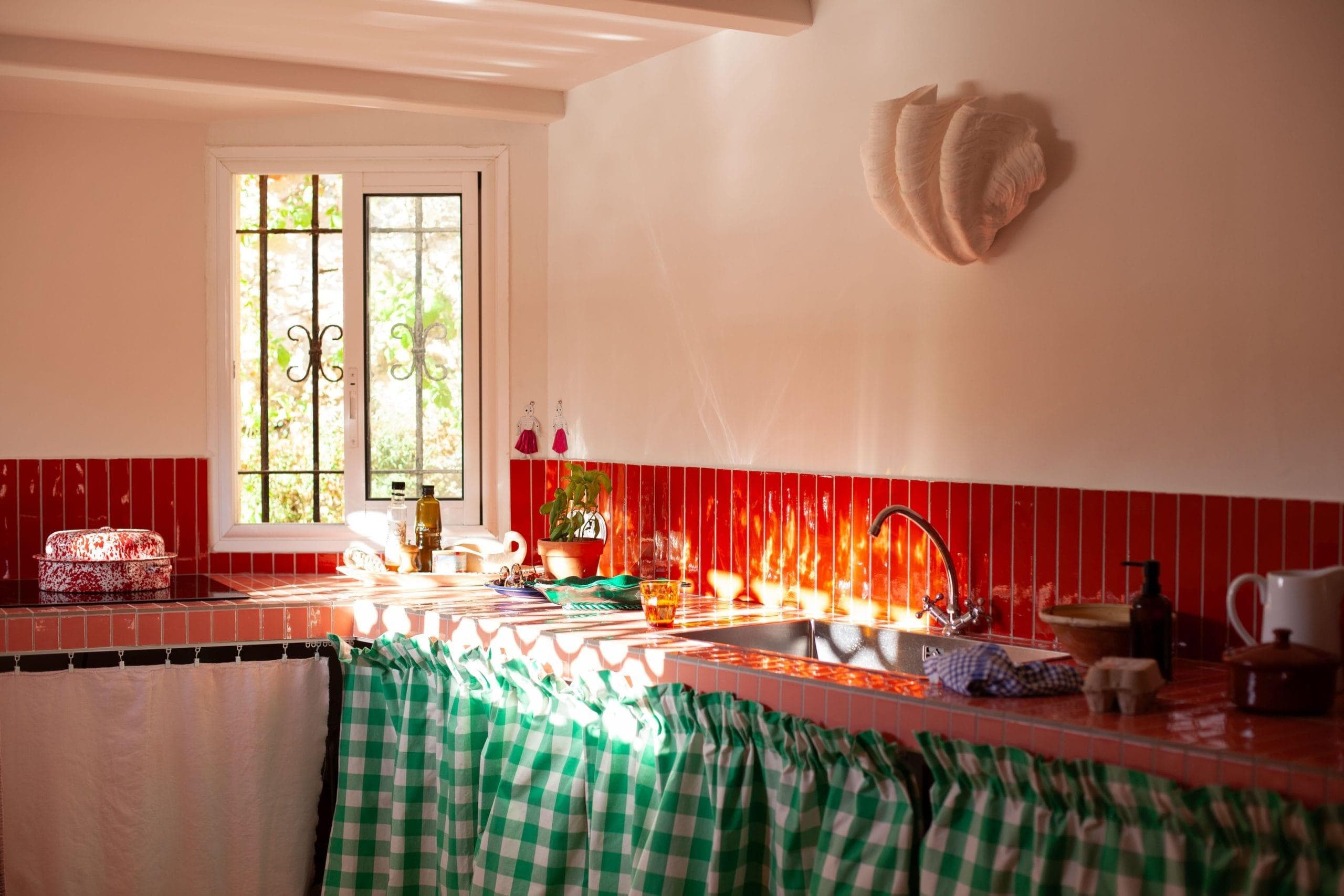 Une cuisine chaleureuse à l'ambiance vintage rappelant le Cabanon des Goudes, avec des murs carrelés de rouge, une lumière du soleil qui entre par la fenêtre et un évier orné d'un rideau à carreaux verts. Un panier, une plante et du liquide vaisselle ornent le plan de travail, renforçant son ambiance chaleureuse.