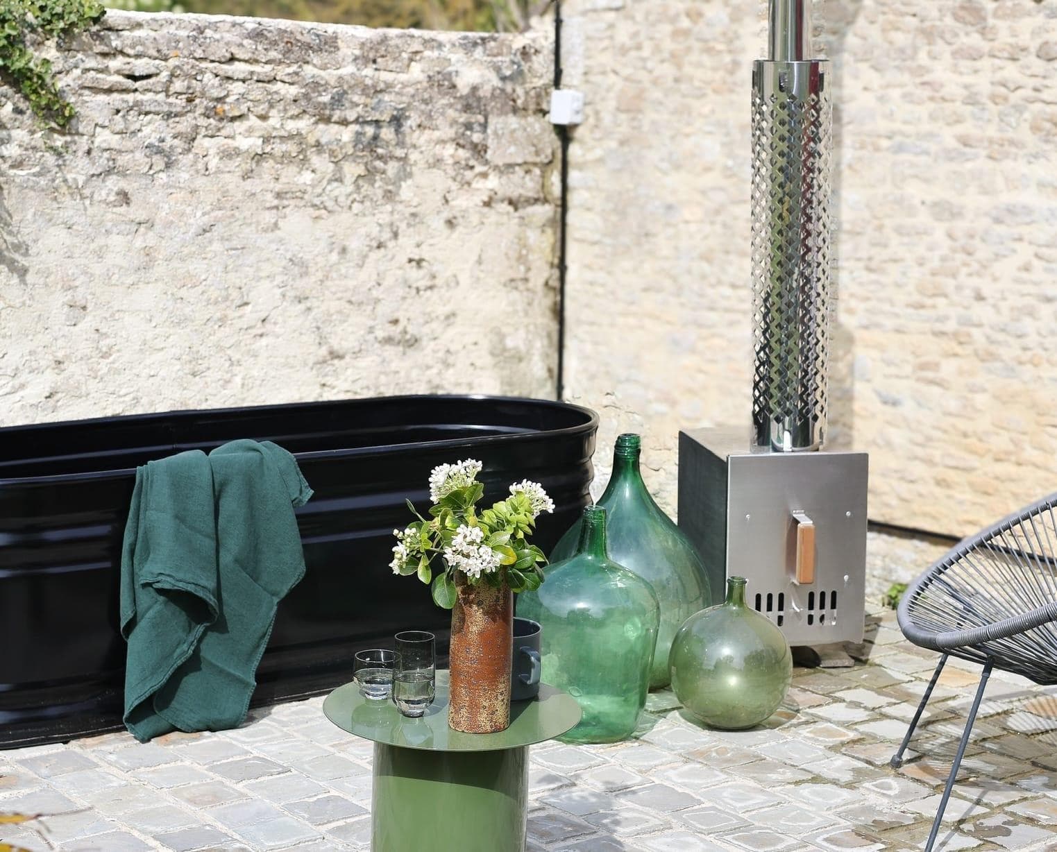 Un patio extérieur moderne avec une baignoire noire, une serviette verte, une petite table verte avec des fleurs et des verres, de grands vases en verre, un radiateur en métal et des murs en pierre grise en arrière-plan.