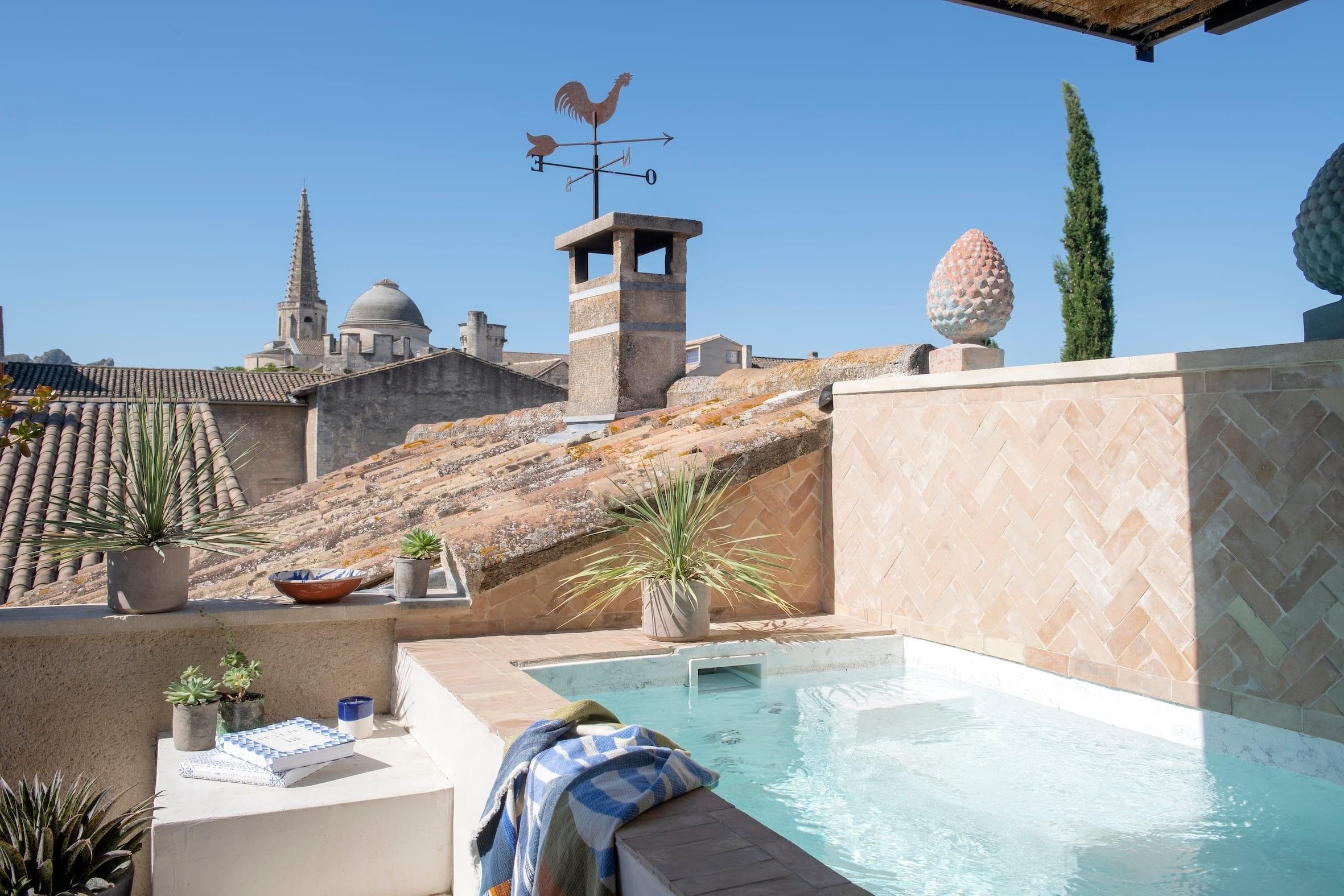 A rooftop terrace featuring a small pool with a towel draped over the edge. There are potted plants around the pool, a weather vane with a rooster on the chimney, and a scenic view of rooftops and a church spire in the background.