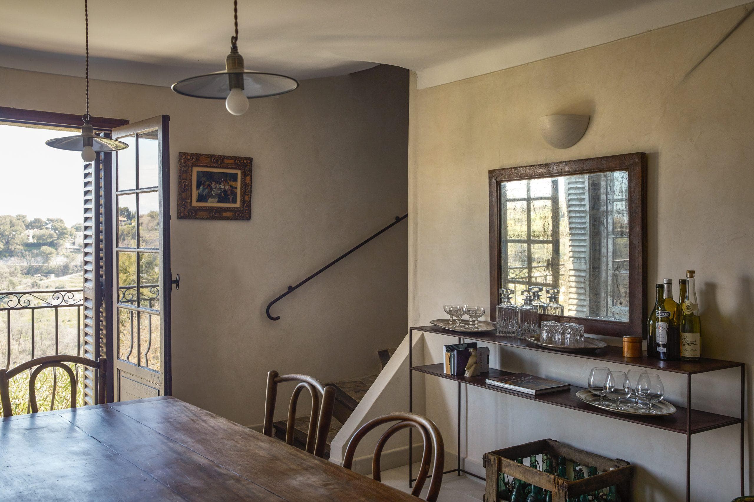 A cozy dining room with a wooden table and chairs. A mirror hangs on the right wall above a sideboard with glasses and bottles. An open door on the left leads to a balcony with a scenic view. A picture frame and a pendant light decorate the space.