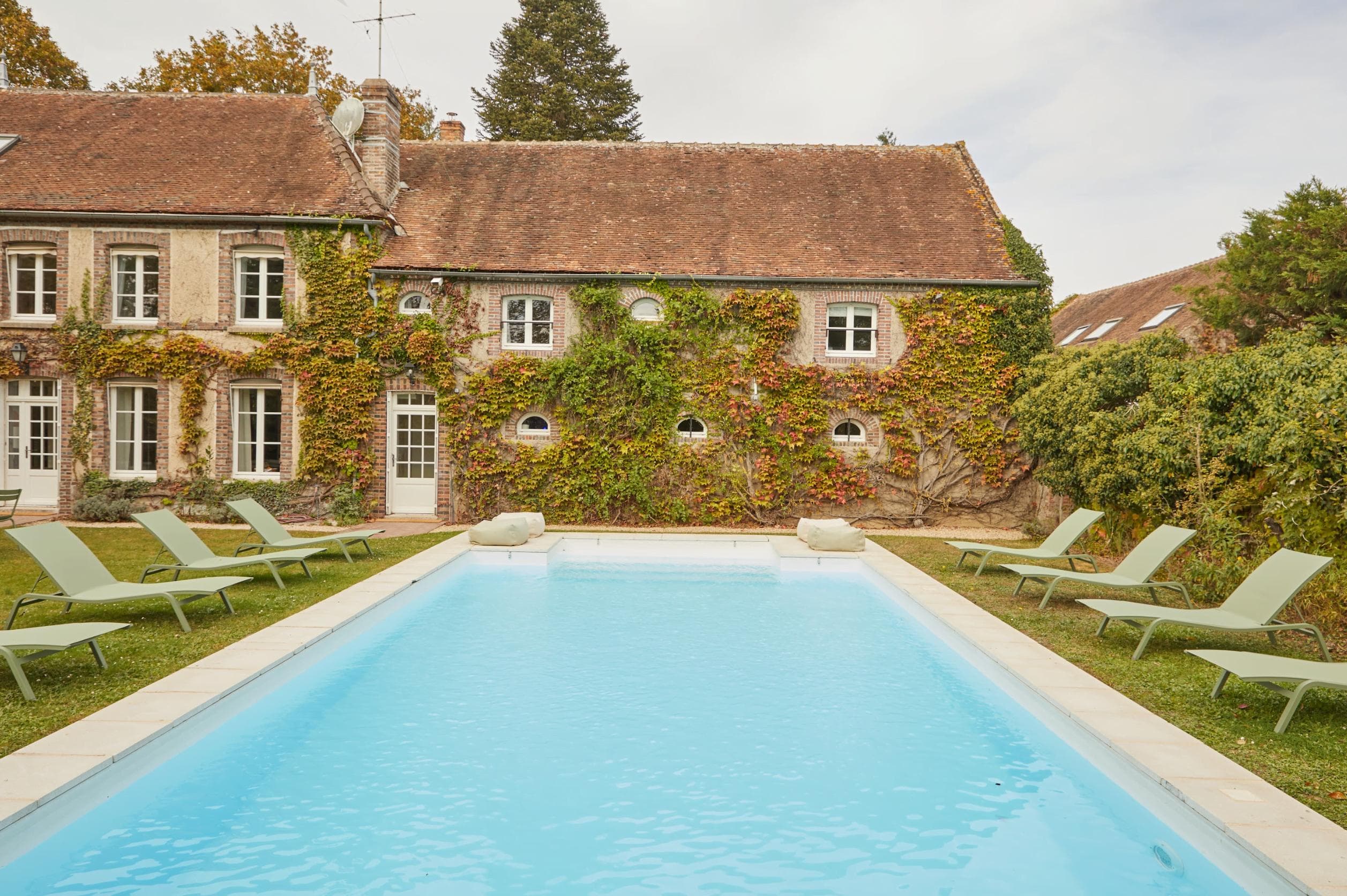 A charming rustic house with ivy-covered walls is seen in the background. In front of the house, there is a bright blue swimming pool surrounded by eight green lounge chairs, set on a well-maintained lawn under a cloudy sky.