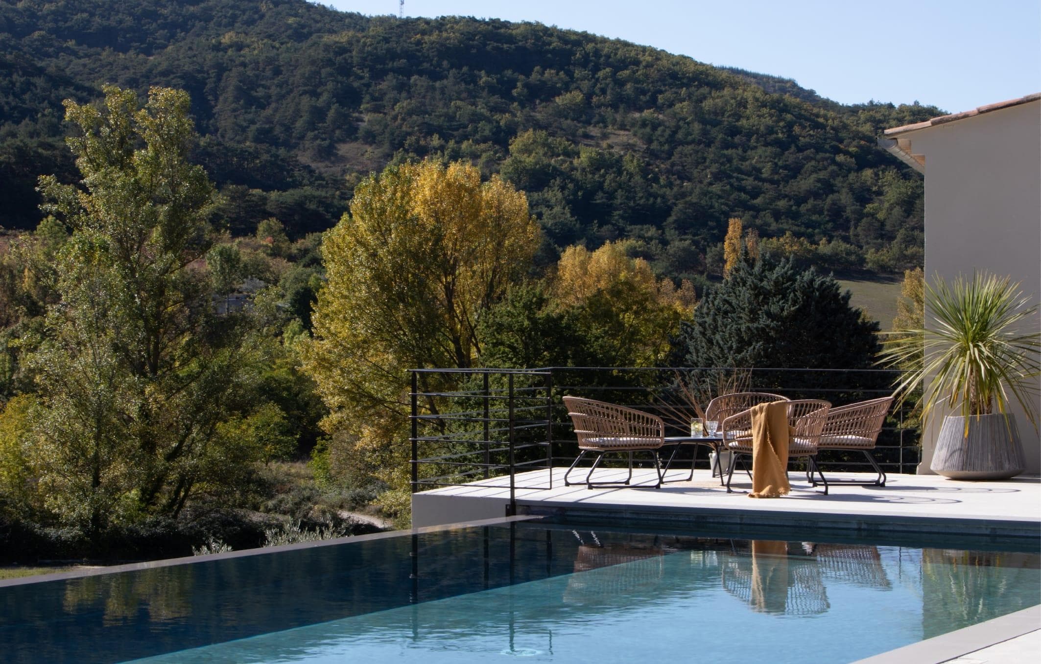 A serene outdoor patio with pool, rattan chairs and table overlooks green hills and trees under a clear sky.