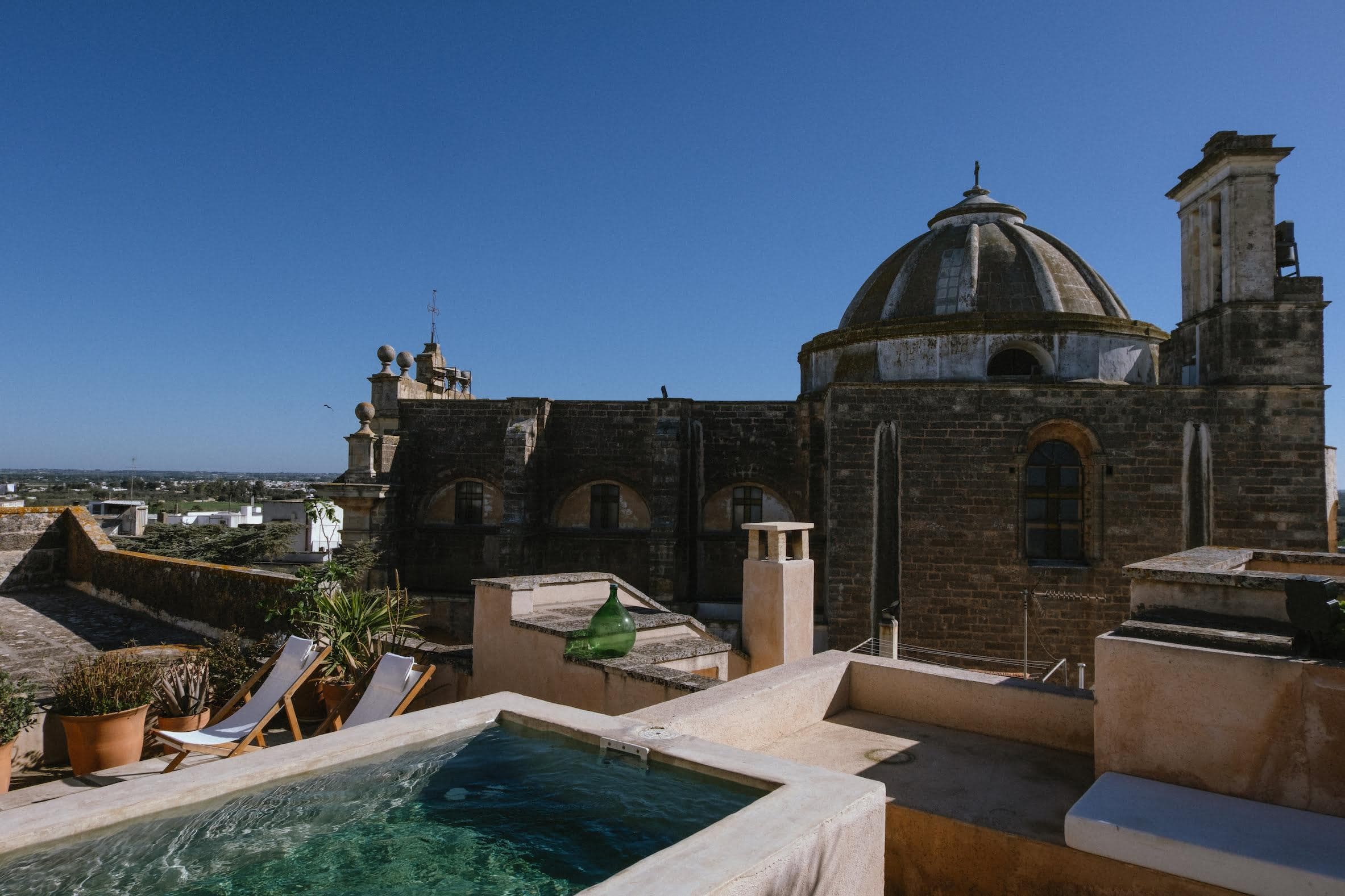 Un toit panoramique avec une petite piscine, des chaises longues et des plantes en pot surplombant une ancienne structure en forme de dôme de pierre sous un ciel bleu clair. La vue s'étend jusqu'aux villas vacances luxe Italie, présentant un paysage lointain de bâtiments blancs et de verdure luxuriante.