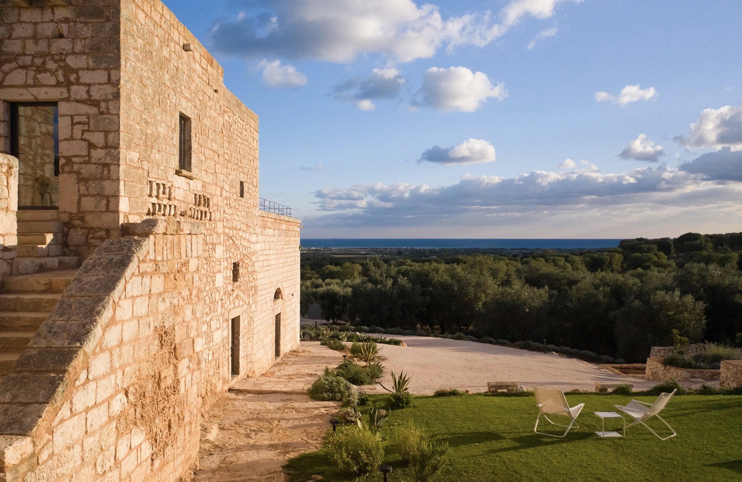 Un bâtiment en pierre se dresse devant un vaste paysage de champs verdoyants et un ciel bleu clair parsemé de nuages. Au premier plan, deux chaises longues blanches sont posées sur une pelouse soigneusement tondue, offrant une vue sereine sur la campagne.