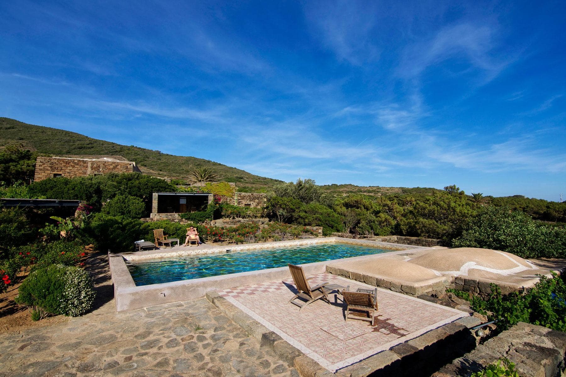 A serene outdoor scene featuring a rectangular swimming pool framed by stone pavement and surrounded by lush greenery. Two wooden lounge chairs sit by the pool, and rolling hills covered with vegetation are visible under a clear blue sky.