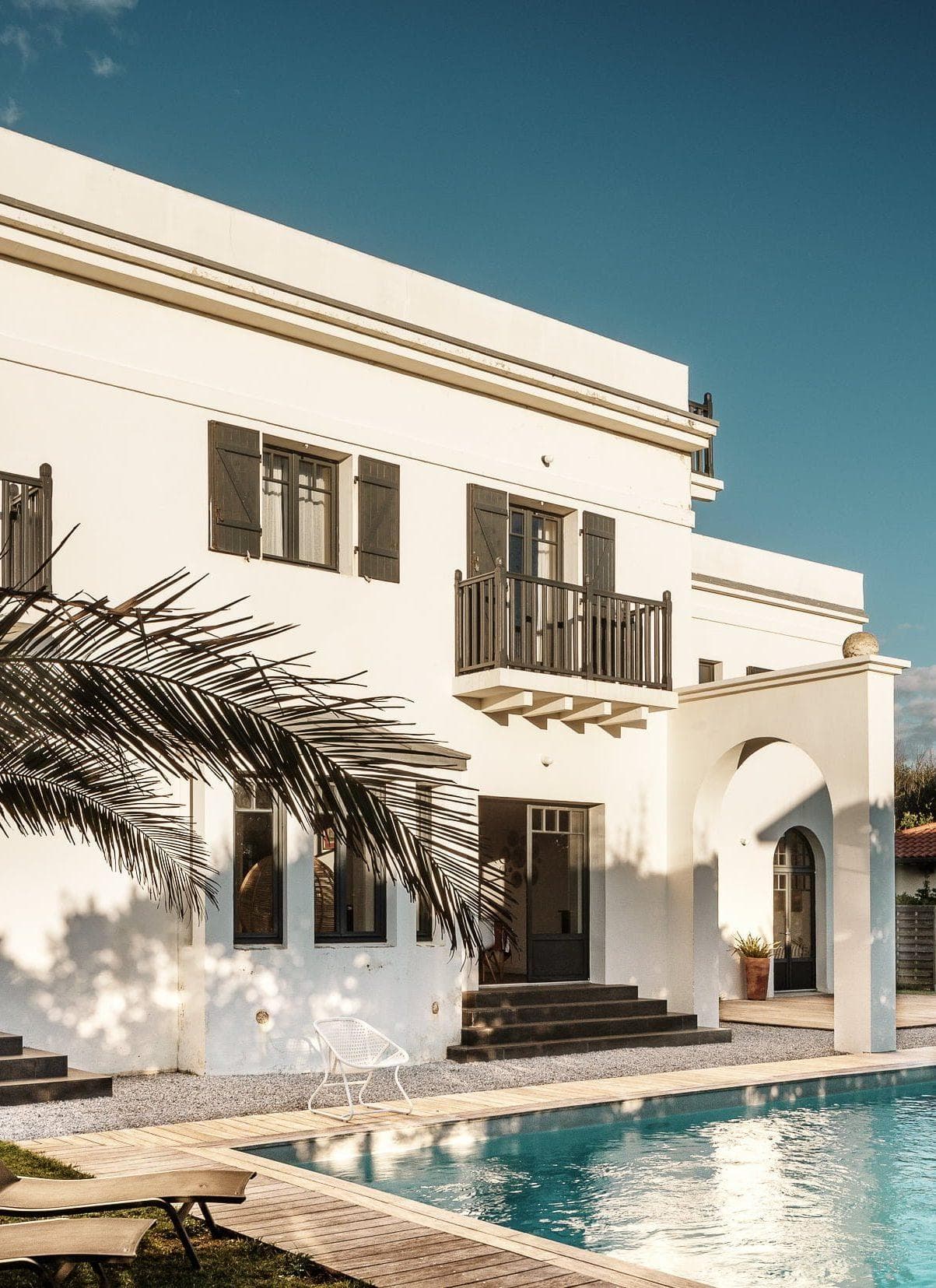 Cette villa de style méditerranéen avec sa façade blanche, ses volets noirs et ses petits balcons offre un lieu de retraite idyllique. Une grande piscine reflète le bâtiment au premier plan, tandis qu'un palmier projette des ombres sur les murs ensoleillés. Deux chaises longues sont installées au bord de la piscine sous un ciel bleu clair, parfait pour une relaxation de yoga.