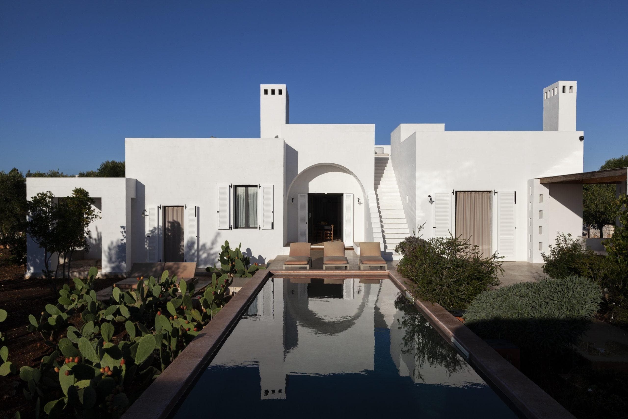 Une villa moderne en stuc blanc aux toits plats et à l'architecture minimaliste. Elle dispose d'une arcade centrale, de plusieurs cheminées et d'un escalier. Une piscine rectangulaire se trouve devant, entourée de verdure, notamment de cactus. Le ciel au-dessus est clair et bleu.