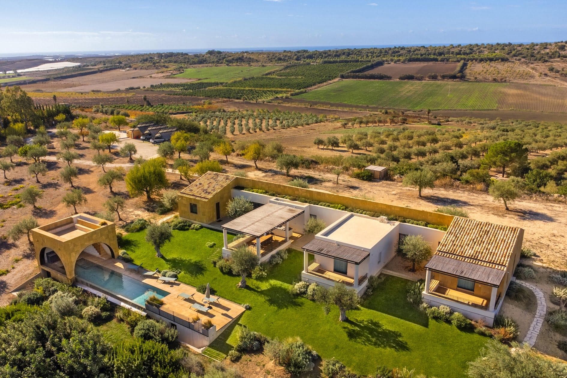 Aerial view of a modern countryside estate with multiple buildings set amidst lush greenery and farmland. The property features a swimming pool with a pergola-covered patio, surrounded by landscaped gardens and olive trees, against a backdrop of vast agricultural fields.