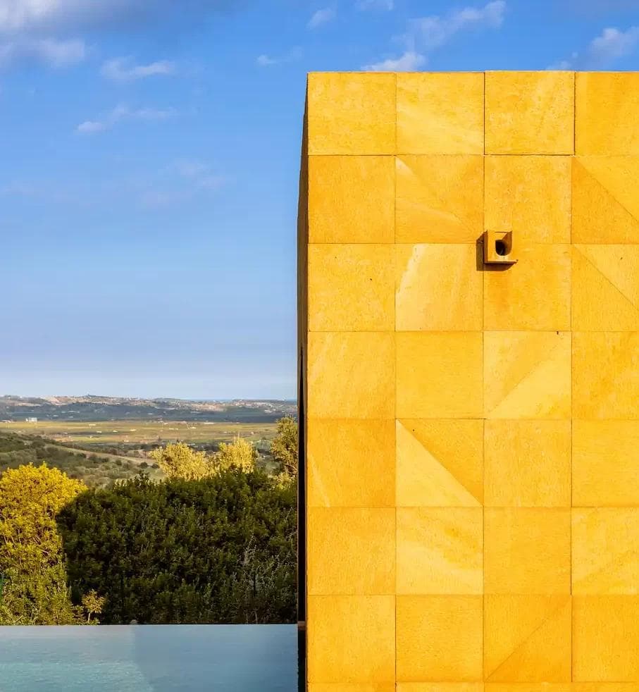 In the foreground stands a geometric, modern building in bright yellow with a surveillance camera mounted on it. Below, an infinity pool blends into the horizon, while lush vegetation and distant hills extend under a blue sky dotted with a few clouds.