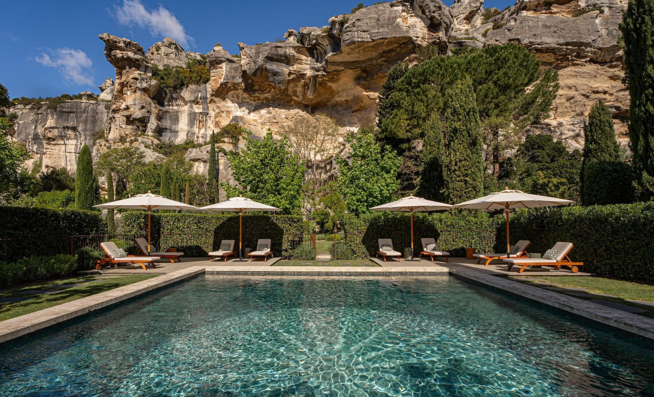 Une luxueuse piscine aux eaux claires, entourée de chaises longues et de parasols, se dresse dans un jardin luxuriant. De hautes falaises et des formations rocheuses se dressent de façon spectaculaire en arrière-plan sous un ciel bleu.