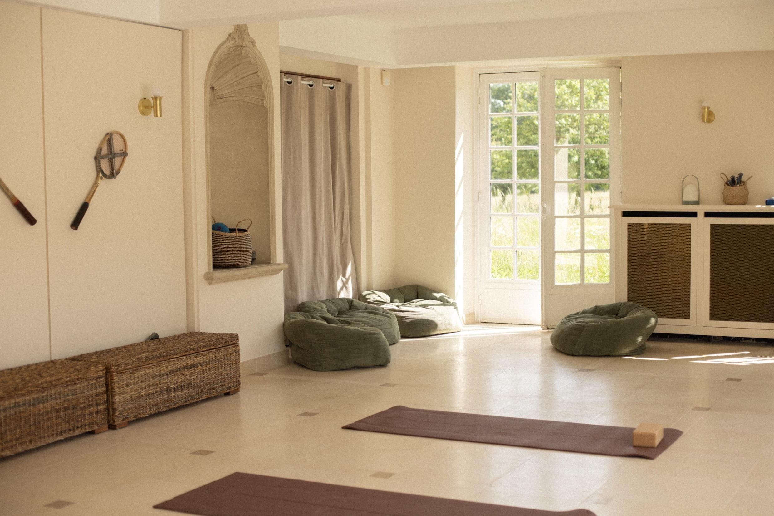 Un studio de yoga serein avec de grandes fenêtres laissant entrer la lumière naturelle. Deux tapis de yoga sont disposés sur le sol, entourés de bancs de rangement en osier, de poufs verts et d'une étagère d'angle avec des accessoires de yoga et un panier tressé. Des raquettes de tennis sont accrochées au mur.