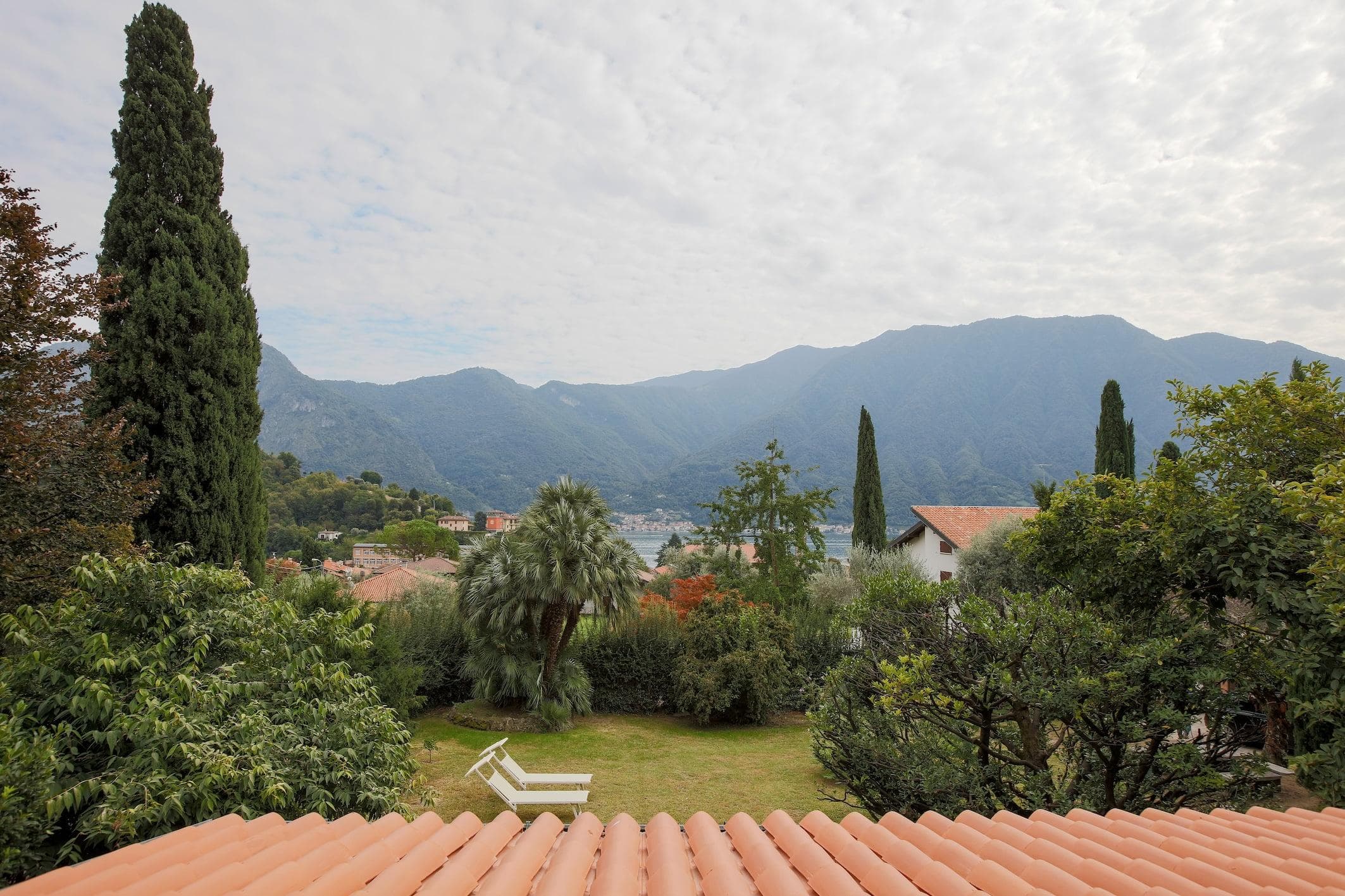 A serene landscape shows a terracotta rooftop in the foreground, leading to a green garden with trees and a white lawn chair. Beyond the garden, a picturesque view of a lake is framed by verdant mountains under a cloudy sky.