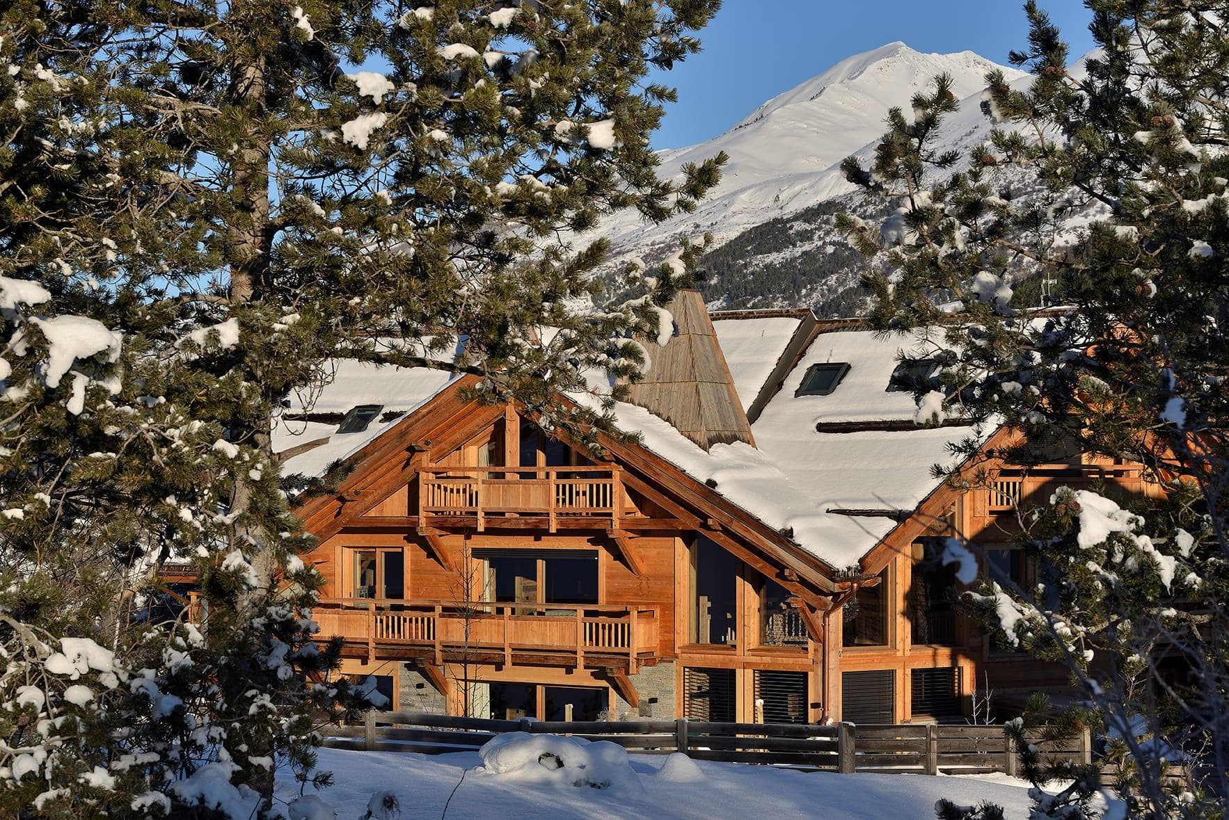 Un chalet en bois confortable avec plusieurs balcons et des toits enneigés est entouré de pins chargés de neige. En arrière-plan, une majestueuse montagne enneigée est visible sur un ciel bleu clair. La scène respire une atmosphère hivernale sereine.