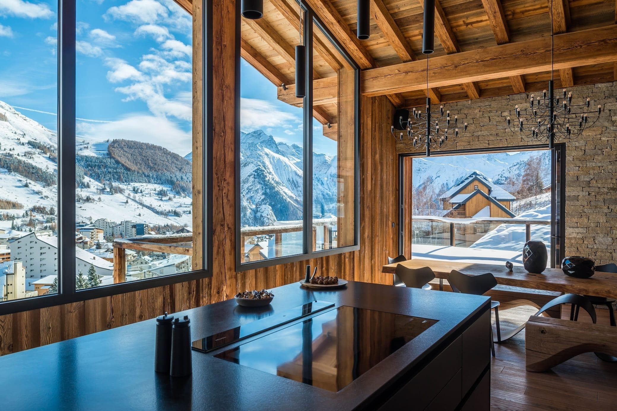 A modern kitchen and dining area with large windows showcasing a snowy mountain view. The interior features wooden walls and ceilings, black countertops, and sleek furnishings. The dining table has black chairs, and the room is filled with natural light.