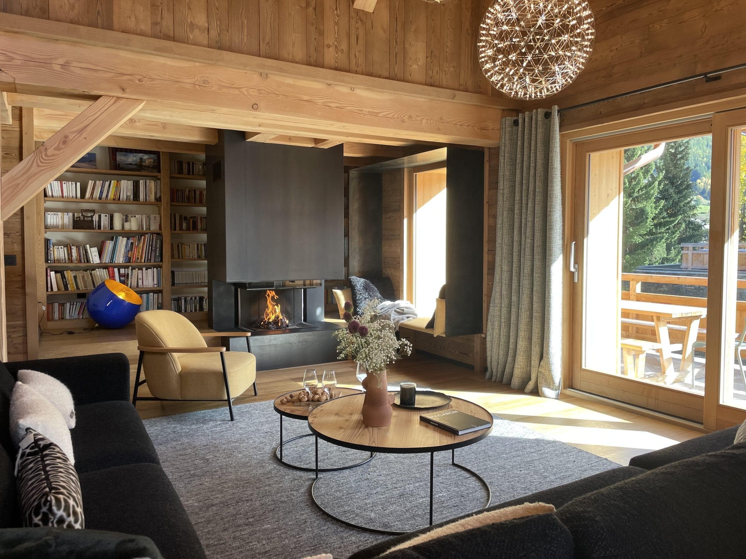 A cozy living room with wooden walls and ceiling. A modern fireplace is flanked by bookshelves. A round chandelier hangs above, and large windows offer a view of trees. The room features a black sofa, beige chair, and circular coffee tables.